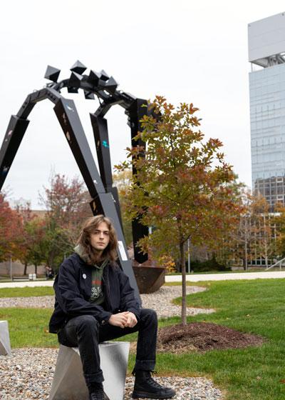 Young male student sitting outside in front of a 20-foot metal sculpture